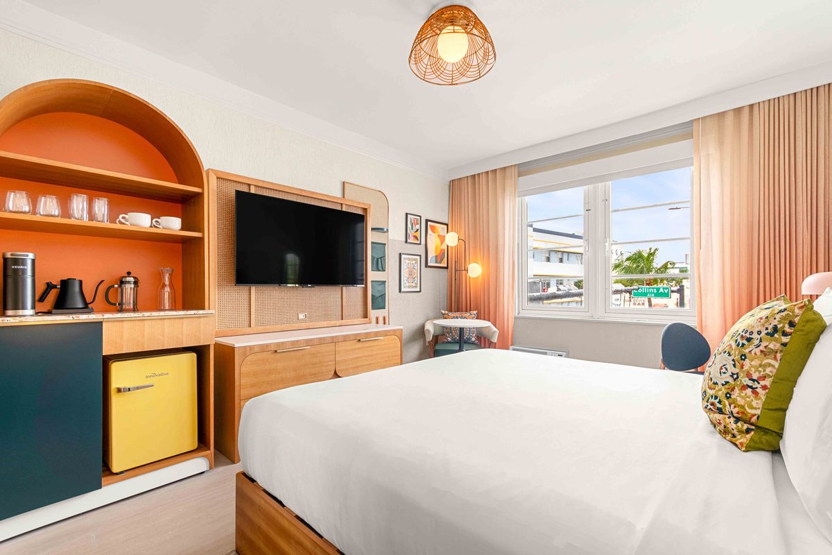 King Suite kitchenette: an arched alcove with glassware and a coffee set, a wall-mounted television and a yellow under-counter fridge, with the bed in the foreground and palms beyond the window