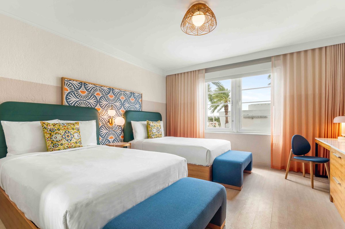 Standard Double Suite: two double beds with white linen against a blue-tiled headboard panel, a blue bench at the foot and a desk with a chair by the window