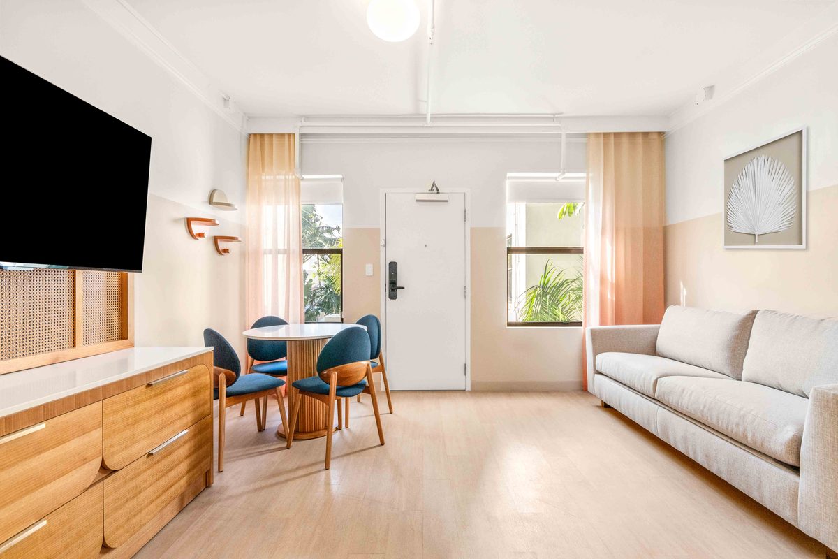 Two-bedroom Villa living area: a large television above a wooden dresser, a round dining table with blue chairs, a cream sofa and a door to the outside