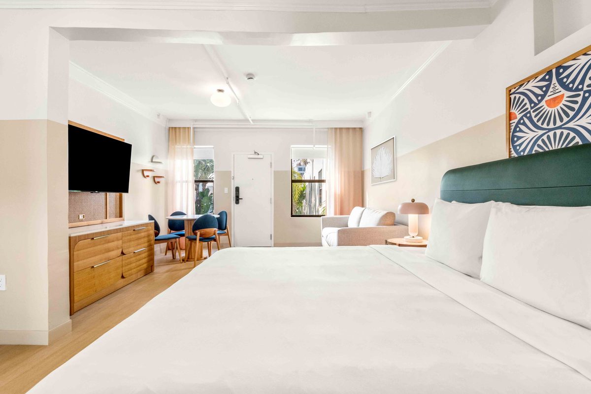 Two-bedroom Villa: a wide open-plan view with a bed in the foreground, the television and dresser at the left, and a dining table with blue chairs beyond