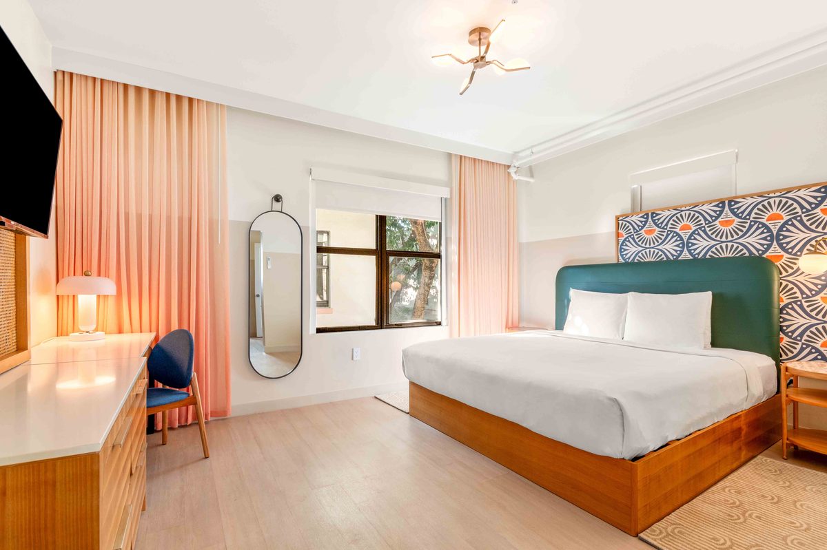 The Villa: an open layout with the kitchen counter in the foreground, a king bed with a teal headboard beyond it, a desk, a tall mirror and windows along the far wall