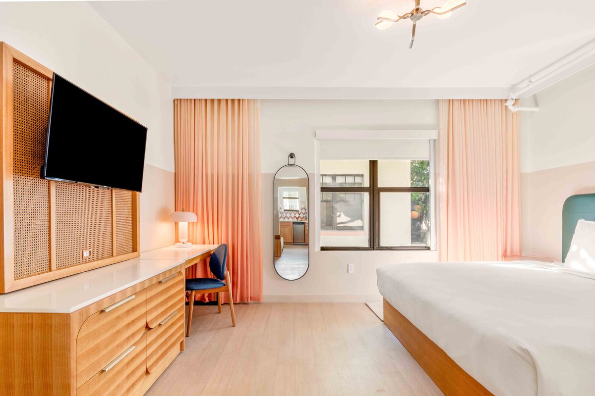 The Villa: a wall-mounted television above a long wooden dresser, an oval mirror, a desk with a blue chair, and the bed to the right under peach curtains